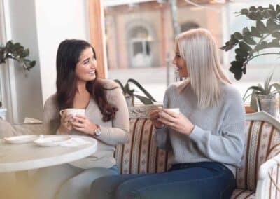 Mainoskuva, jossa kaksi naista istuu kahvilassa ja pitelee kahvikuppeja käsissään.